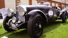 A vintage black classic car with prominent circular headlights and chrome details is parked on a lush green lawn. It features a distinct front grille, large tires, and a 'MotoClassic' license plate. The surroundings include a brick building and various other vehicles.