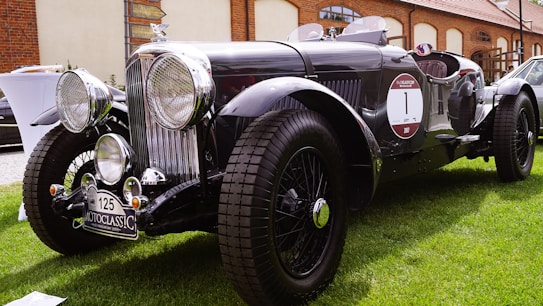 A vintage black classic car with prominent circular headlights and chrome details is parked on a lush green lawn. It features a distinct front grille, large tires, and a 'MotoClassic' license plate. The surroundings include a brick building and various other vehicles.