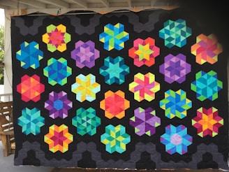 A quilt featuring a pattern of geometric star-shaped designs in a variety of bright colors, including blue, green, yellow, pink, and purple, set against a black background. The quilt appears to be hanging outside, possibly on a porch.