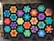 A quilt featuring a pattern of geometric star-shaped designs in a variety of bright colors, including blue, green, yellow, pink, and purple, set against a black background. The quilt appears to be hanging outside, possibly on a porch.