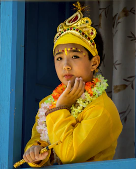A child wearing an elegant sharara set in soft cream tones, seated on a traditional wooden swing decorated with marigold garlands.