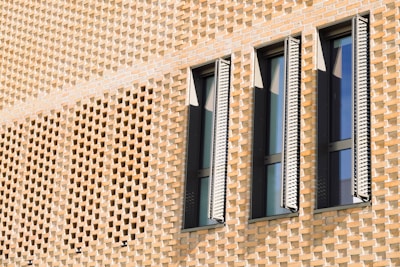 A detailed pattern of light brown bricks forms a textured wall. Three narrow, vertical windows with slatted shutters are spaced evenly across, casting distinct shadows onto the surface. The structure combines traditional architectural elements with a modern aesthetic, highlighting intricate brickwork and contemporary window design.