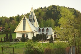 A modern church with sharp angular architecture features prominently in a lush green landscape. The building has striking triangular rooflines and a tall, pointed steeple. Surrounding the church, neatly manicured trees and shrubs are visible, with a dense forest forming a natural background. The sky is overcast, adding a serene feel to the environment.
