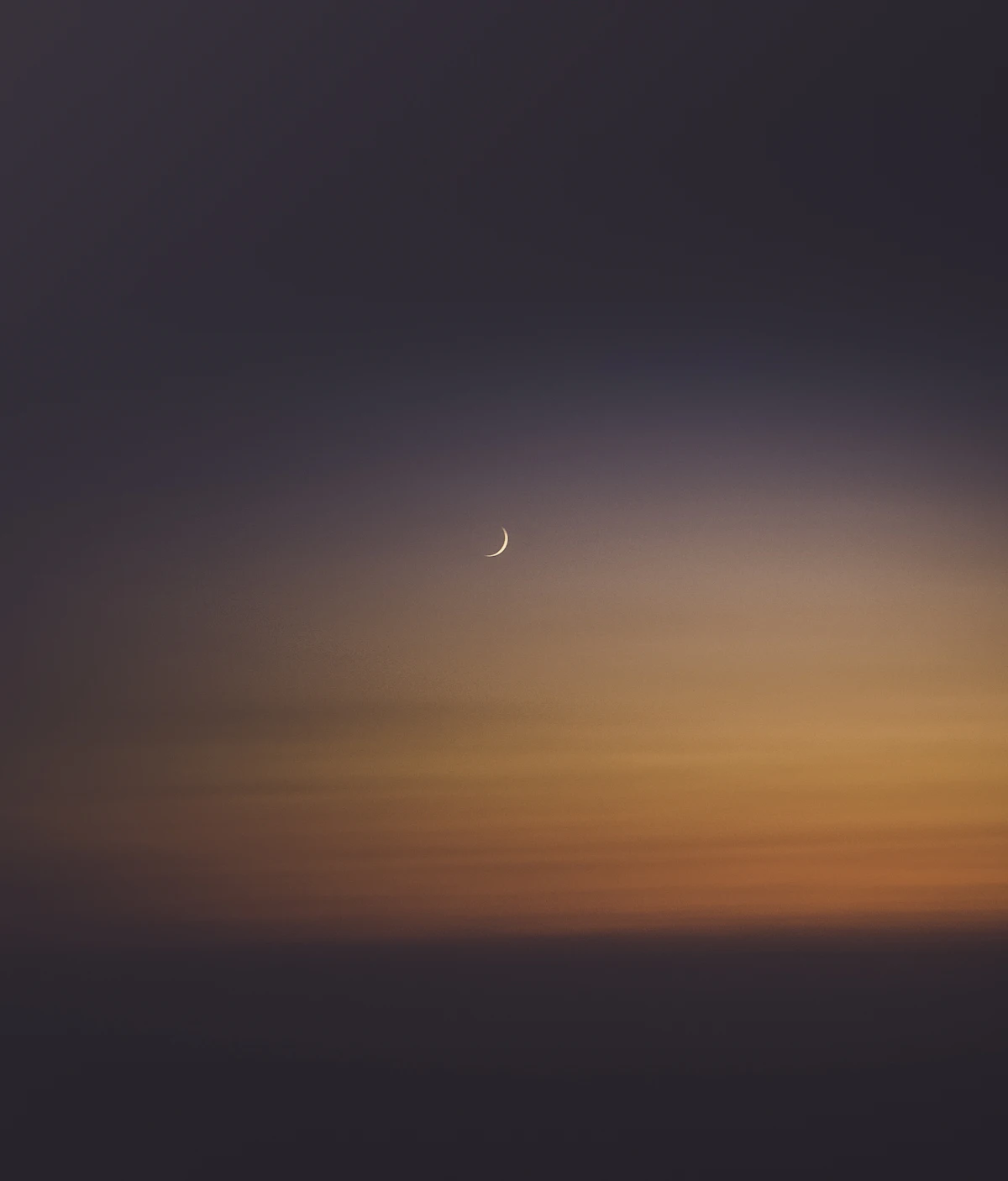 Fin croissant de lune dans un ciel de crépuscule aux tons or et crème, au-dessus d'un horizon marin