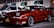 A red Toyota sports car is parked in a dimly lit street next to a white Ford vehicle. The scene includes several buildings in the background with green awnings and visible business signs. The cars are well-maintained and appear to be in a commercial area.