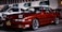 A red Toyota sports car is parked in a dimly lit street next to a white Ford vehicle. The scene includes several buildings in the background with green awnings and visible business signs. The cars are well-maintained and appear to be in a commercial area.