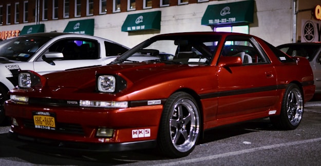 A red Toyota sports car is parked in a dimly lit street next to a white Ford vehicle. The scene includes several buildings in the background with green awnings and visible business signs. The cars are well-maintained and appear to be in a commercial area.