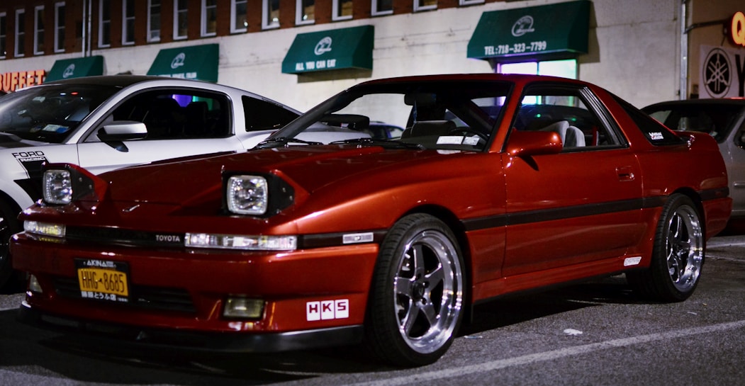 A red Toyota sports car is parked in a dimly lit street next to a white Ford vehicle. The scene includes several buildings in the background with green awnings and visible business signs. The cars are well-maintained and appear to be in a commercial area.