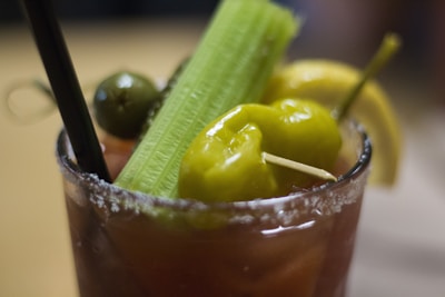 A cocktail glass filled with a beverage that has a salted rim. The drink is garnished with a celery stalk, green olives, a yellow pepper, and a lemon wedge. A black straw is also present in the glass.