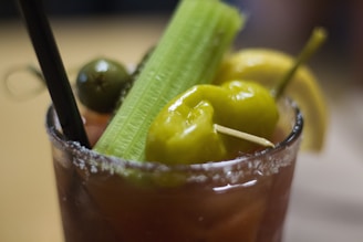 A cocktail glass filled with a beverage that has a salted rim. The drink is garnished with a celery stalk, green olives, a yellow pepper, and a lemon wedge. A black straw is also present in the glass.