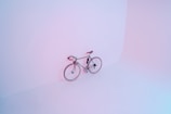 A bicycle is positioned against a minimalist, softly lit wall that blends pastel pink and blue hues. The scene is clean and simple, emphasizing the streamlined design of the bicycle.