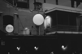 A monochrome image of an urban building with geometric shapes and large spherical light fixtures casting a soft glow. The facade features different textures and architectural elements, including beams, windows, and pipes. The word 'OGG' is visible on one of the spheres.