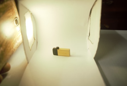 A small, golden USB flash drive is placed in a makeshift white lightbox. The setup includes a light source on the left and white walls surrounding the USB stick, which is positioned centrally.