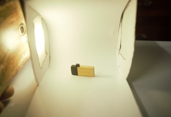 A small, golden USB flash drive is placed in a makeshift white lightbox. The setup includes a light source on the left and white walls surrounding the USB stick, which is positioned centrally.