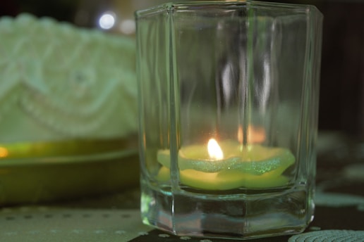 A tealight candle placed inside a square glass holder emits a warm glow, casting subtle reflections on the glass surfaces. The candle is situated on a patterned fabric background, and a soft-focus decorative element with intricate designs is visible in the background.