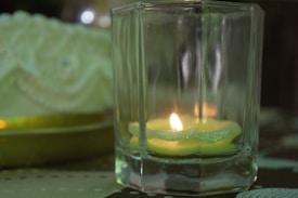 A tealight candle placed inside a square glass holder emits a warm glow, casting subtle reflections on the glass surfaces. The candle is situated on a patterned fabric background, and a soft-focus decorative element with intricate designs is visible in the background.