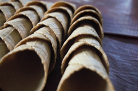 A close-up view of several stacked ice cream cones, arranged in neat rows. The cones have a textured, waffle pattern and are positioned on a dark surface, suggesting a tabletop or counter.
