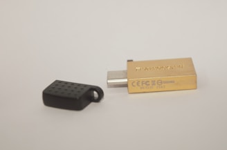 A USB flash drive with a metallic gold casing lies next to its black cap on a plain background. The cap has a textured surface.