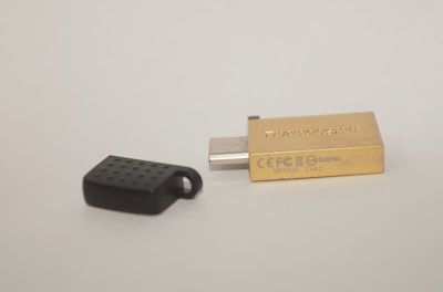A USB flash drive with a metallic gold casing lies next to its black cap on a plain background. The cap has a textured surface.