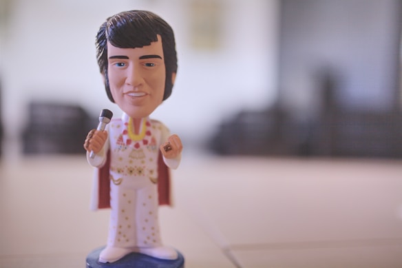 A stylized figurine dressed in a white jumpsuit with gold and red decorations, holding a microphone. The figurine has dark hair and stands on a small blue base against a blurred background.