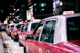A line of red taxis is parked along a city street at night, with bright neon lights reflecting on their surfaces. The taxis are waiting in a queue, and the background is slightly blurred, showcasing the vibrant city life.