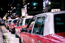 A line of red taxis is parked along a city street at night, with bright neon lights reflecting on their surfaces. The taxis are waiting in a queue, and the background is slightly blurred, showcasing the vibrant city life.