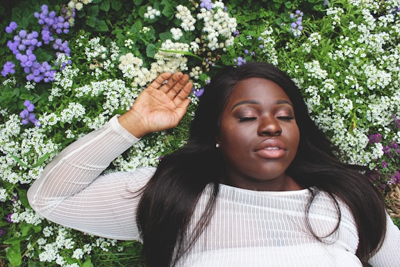 A woman with closed eyes lies peacefully on a bed of green foliage and small, white, and purple flowers, wearing a sheer white top. Her relaxed expression and surroundings suggest tranquility and nature.