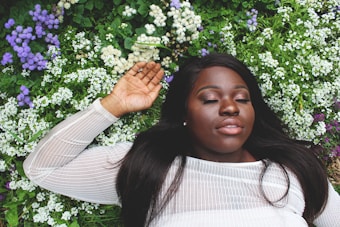 A woman with closed eyes lies peacefully on a bed of green foliage and small, white, and purple flowers, wearing a sheer white top. Her relaxed expression and surroundings suggest tranquility and nature.