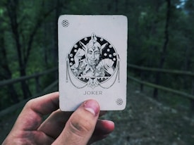 A hand holding a playing card with a 'Joker' illustration. The card features an intricate black and white design of a jester character holding a fan of cards. The background is a blurred outdoor scene with trees.