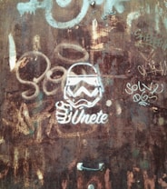 A rusty metal surface with various graffiti, including a stylized image of a helmeted figure, resembling a stormtrooper, accompanied by the text '&Uacute;nete' in bold white lettering. Additional graffiti is visible, including the words 'Solve' and 'DGS' in different lettering styles.
