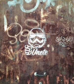 A rusty metal surface with various graffiti, including a stylized image of a helmeted figure, resembling a stormtrooper, accompanied by the text 'Únete' in bold white lettering. Additional graffiti is visible, including the words 'Solve' and 'DGS' in different lettering styles.