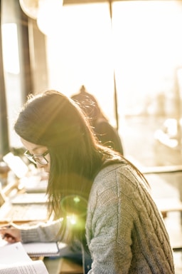 A person with long hair and glasses, wearing a cozy knitted sweater, studies at a desk. The setting is bright with sunlight streaming through a large window, illuminating the scene with warm and soft light. Papers and a laptop are visible, indicating a study or work environment.
