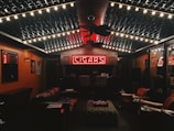 A cozy, dimly lit room featuring a neon sign that reads 'CIGARS' at the far wall. The ceiling is decorated with string lights, creating a warm ambiance. Leather chairs and sofas are arranged around a central ottoman, and the walls are lined with wood paneling and display cases.