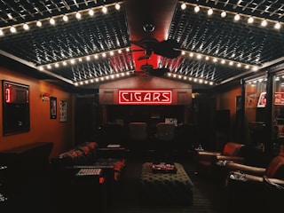 A cozy, dimly lit room featuring a neon sign that reads 'CIGARS' at the far wall. The ceiling is decorated with string lights, creating a warm ambiance. Leather chairs and sofas are arranged around a central ottoman, and the walls are lined with wood paneling and display cases.