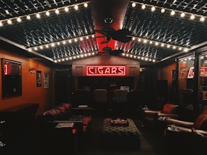 A cozy, dimly lit room featuring a neon sign that reads 'CIGARS' at the far wall. The ceiling is decorated with string lights, creating a warm ambiance. Leather chairs and sofas are arranged around a central ottoman, and the walls are lined with wood paneling and display cases.