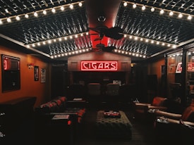 A cozy, dimly lit room featuring a neon sign that reads 'CIGARS' at the far wall. The ceiling is decorated with string lights, creating a warm ambiance. Leather chairs and sofas are arranged around a central ottoman, and the walls are lined with wood paneling and display cases.