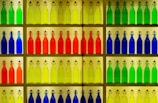 A display of colorful glass bottles arranged neatly on shelves. The bottles are organized by color, including shades of green, yellow, orange, red, and blue. Each bottle has a metal clasp top and a rectangular label. The setup creates a vibrant and visually pleasing pattern.