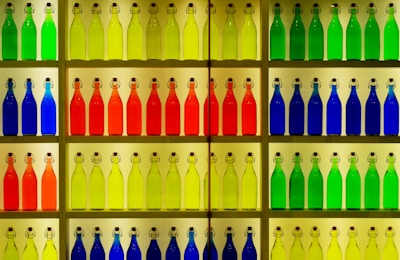 A display of colorful glass bottles arranged neatly on shelves. The bottles are organized by color, including shades of green, yellow, orange, red, and blue. Each bottle has a metal clasp top and a rectangular label. The setup creates a vibrant and visually pleasing pattern.