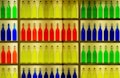 A display of colorful glass bottles arranged neatly on shelves. The bottles are organized by color, including shades of green, yellow, orange, red, and blue. Each bottle has a metal clasp top and a rectangular label. The setup creates a vibrant and visually pleasing pattern.