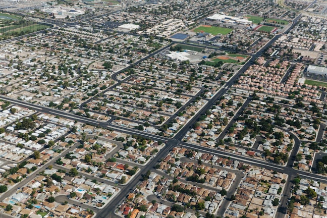 Aerial view of a residential neighborhood with a grid pattern of streets and numerous houses. The area is densely packed with homes featuring various roof colors and some outdoor pools. Trees are scattered throughout, providing greenery between the buildings. At the top, sports fields and larger structures are visible, suggesting a school or community area.