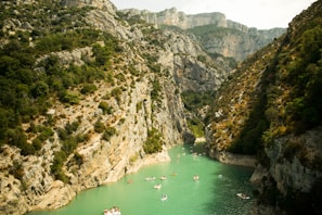 A picturesque canyon with steep rocky cliffs covered in greenery on both sides, and a narrow, turquoise river flowing between them. Several small boats with people are seen leisurely floating on the water, enjoying the scenic surroundings.