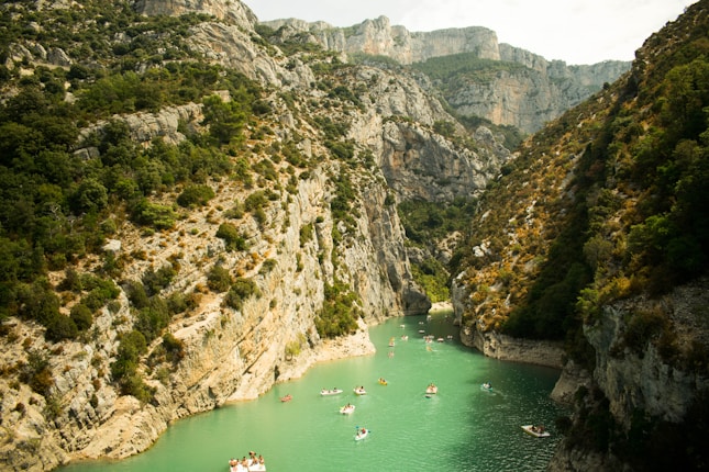 A picturesque canyon with steep rocky cliffs covered in greenery on both sides, and a narrow, turquoise river flowing between them. Several small boats with people are seen leisurely floating on the water, enjoying the scenic surroundings.