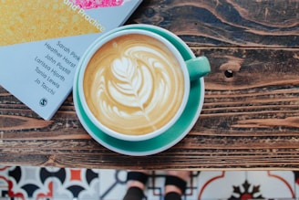 A cup of latte with a leaf-shaped foam art sits on a wooden table. Next to the cup is a book with colorful cover details, showing names of authors. The table has a rustic appearance, and part of a patterned tile floor is visible.