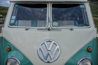 A vintage Volkswagen van with a turquoise and off-white color scheme, featuring a prominent Volkswagen logo on the front. Inside the windshield, a small plush toy is visible on the dashboard.