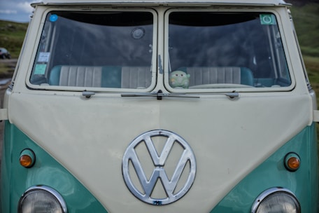 A vintage Volkswagen van with a turquoise and off-white color scheme, featuring a prominent Volkswagen logo on the front. Inside the windshield, a small plush toy is visible on the dashboard.