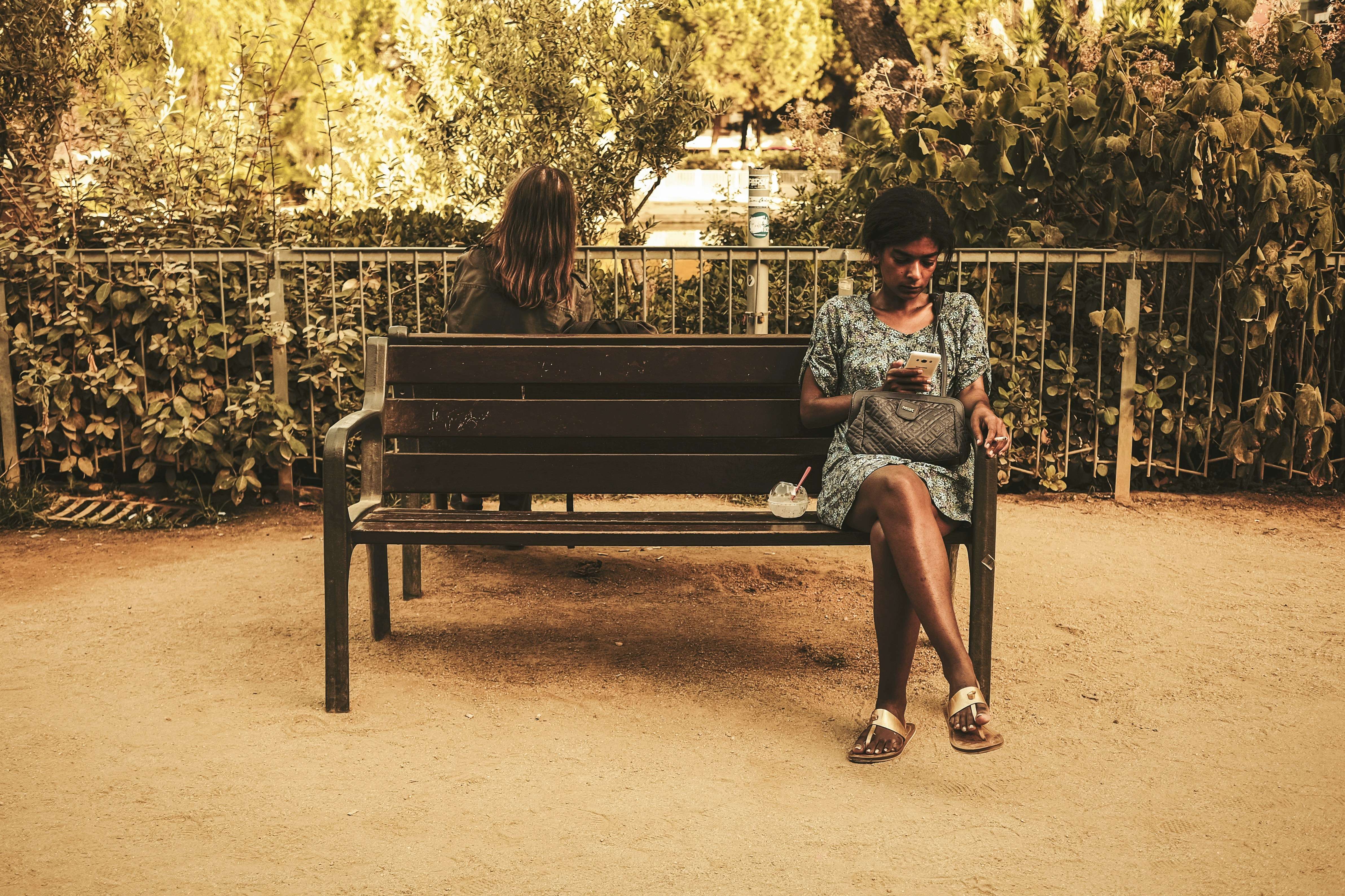 women sitting on a bench. the woman in front is a woman of color, on her phone, smoking a cigarette