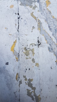A close-up view of a textured wall surface with peeling paint. The surface shows various shades of gray, with patches of yellow and black. The texture appears rough and cracked, giving an aged and weathered appearance.