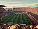 A large football stadium filled with spectators watching a match. The green pitch is lined with players in action, surrounded by packed stands. Banners and advertisements are visible around the stadium.