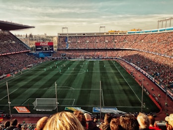 A large football stadium filled with spectators watching a match. The green pitch is lined with players in action, surrounded by packed stands. Banners and advertisements are visible around the stadium.