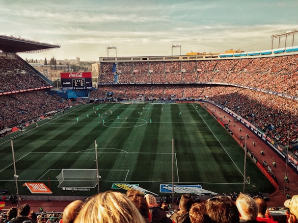 A large football stadium filled with spectators watching a match. The green pitch is lined with players in action, surrounded by packed stands. Banners and advertisements are visible around the stadium.